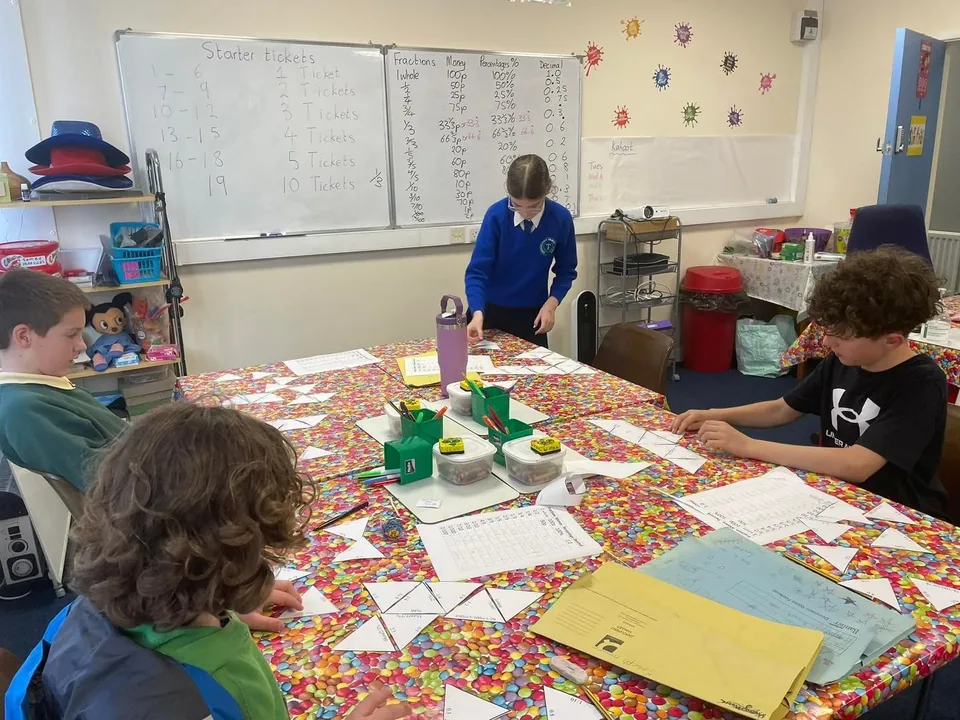A Sumkids maths class in Limavady, with pupils working at a colourful table and a whiteboard of fractions, decimals and percentages
